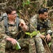 Two military personnel in MultiCam/OCP camouflage uniforms are seated on a hillside in a jungle or forested environment. The soldier on the left appears to be briefing or explaining something while holding a small notebook and gesturing with his hand. Both carry tactical gear including radios and backpacks, and wear US flag patches on their shoulders.
