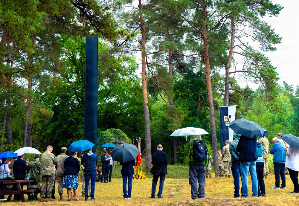 An outdoor memorial or commemoration ceremony is taking place in a forested area during rainy weather. Attendees, including military personnel in camouflage uniforms and civilians, are gathered around a tall dark monolithic monument or obelisk. A banner or flag with a coat of arms is visible, suggesting an official or governmental event.
