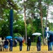 An outdoor memorial or commemoration ceremony is taking place in a forested area during rainy weather. Attendees, including military personnel in camouflage uniforms and civilians, are gathered around a tall dark monolithic monument or obelisk. A banner or flag with a coat of arms is visible, suggesting an official or governmental event.