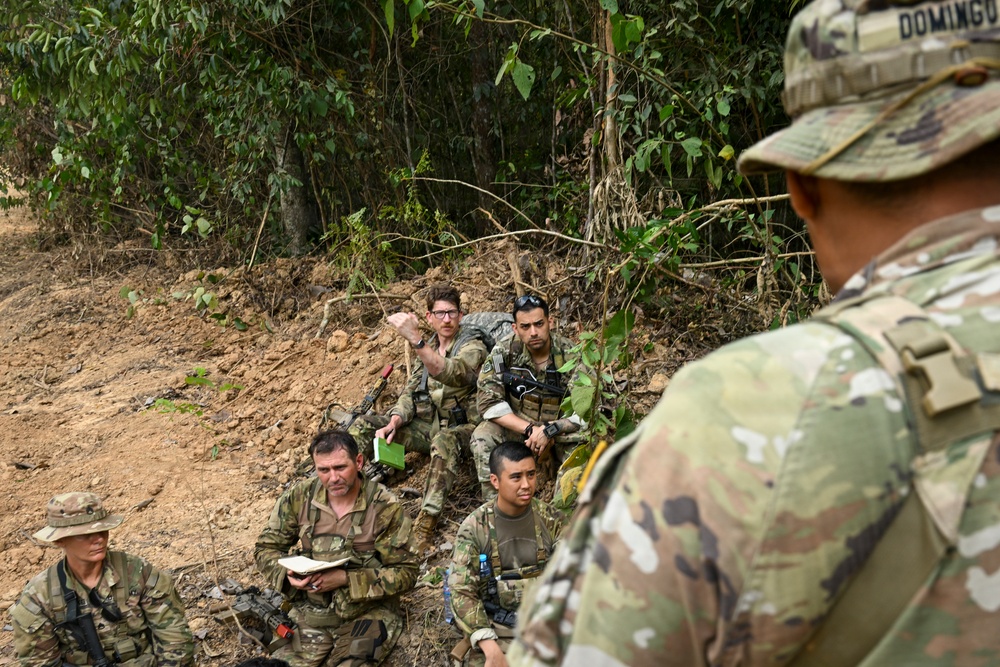 A group of military personnel in camouflage uniforms are gathered in a jungle or forested terrain, appearing to receive a briefing or instruction. One soldier in the foreground, identified by a name tape reading 'DOMINGUEZ,' faces the seated group. The soldiers are equipped with weapons, tactical gear, and at least one is taking notes.