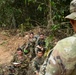 A group of military personnel in camouflage uniforms are gathered in a jungle or forested terrain, appearing to receive a briefing or instruction. One soldier in the foreground, identified by a name tape reading 'DOMINGUEZ,' faces the seated group. The soldiers are equipped with weapons, tactical gear, and at least one is taking notes.