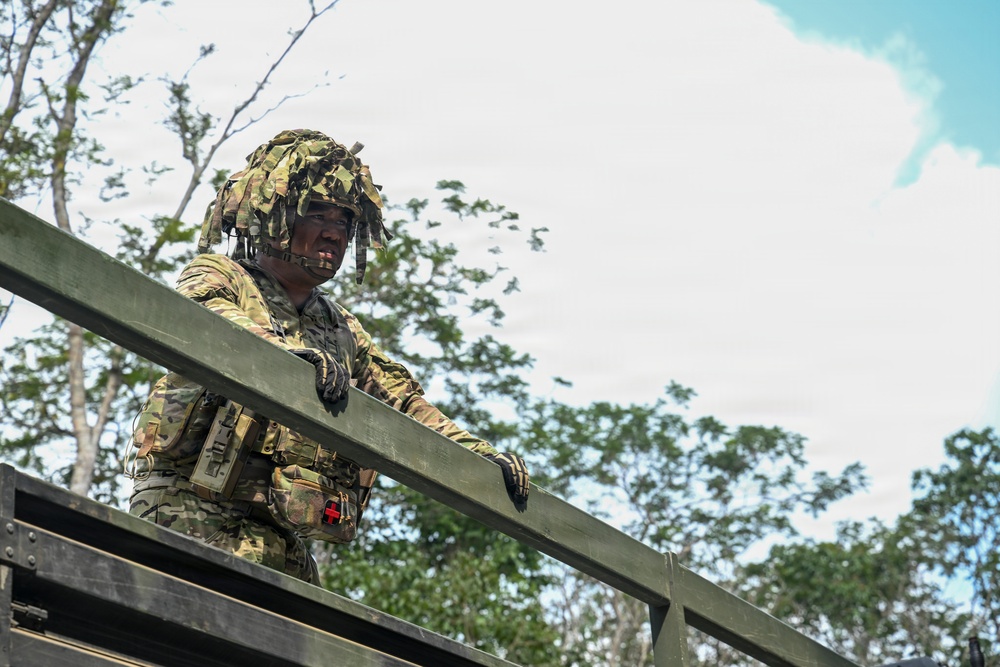 A soldier in full camouflage gear, including a ghillie-style helmet cover, is positioned at an elevated structure in a wooded outdoor environment. The soldier is wearing multicam/OCP pattern uniform with tactical vest, gloves, and a communications headset. The image is taken from a low angle, emphasizing the soldier's elevated position against a partly cloudy sky.