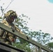 A soldier in full camouflage gear, including a ghillie-style helmet cover, is positioned at an elevated structure in a wooded outdoor environment. The soldier is wearing multicam/OCP pattern uniform with tactical vest, gloves, and a communications headset. The image is taken from a low angle, emphasizing the soldier's elevated position against a partly cloudy sky.