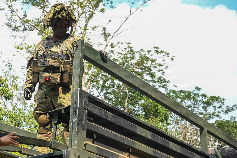 A soldier in full combat gear stands atop a military vehicle, likely a truck or transport, in a tropical or jungle environment. The soldier is wearing multicam camouflage uniform, a helmet with camouflage netting, tactical vest with magazine pouches, and a red cross medical pouch. The image is taken from a low angle, emphasizing the soldier's elevated position against a partly cloudy sky.