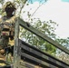 A soldier in full combat gear stands atop a military vehicle, likely a truck or transport, in a tropical or jungle environment. The soldier is wearing multicam camouflage uniform, a helmet with camouflage netting, tactical vest with magazine pouches, and a red cross medical pouch. The image is taken from a low angle, emphasizing the soldier's elevated position against a partly cloudy sky.