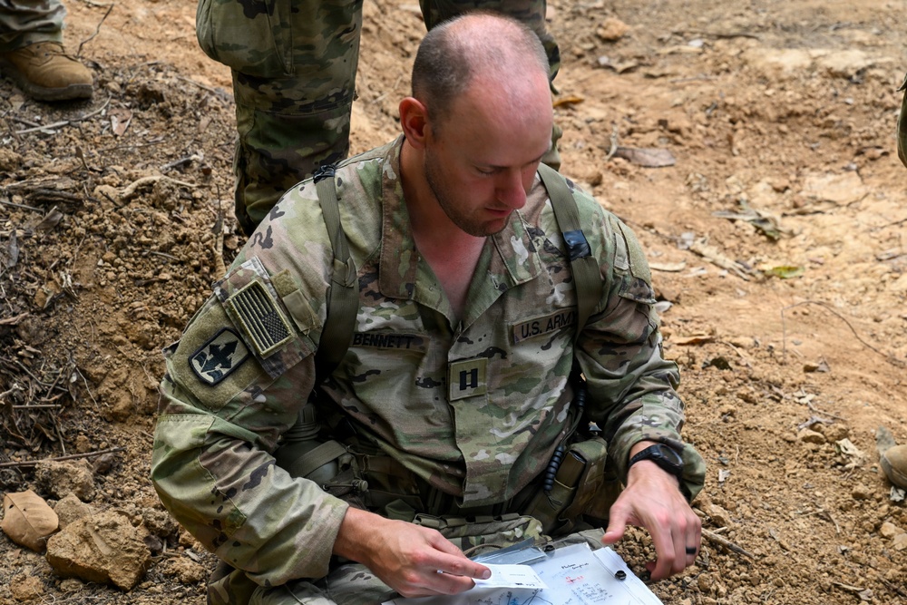A U.S. Army Captain named Bennett is seated on the ground in a field environment, reviewing documents or maps. He is wearing Operational Camouflage Pattern (OCP) uniform with visible rank insignia (Captain's bars) and a unit patch. The setting appears to be an outdoor training or operational area with disturbed soil terrain.