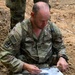 A U.S. Army Captain named Bennett is seated on the ground in a field environment, reviewing documents or maps. He is wearing Operational Camouflage Pattern (OCP) uniform with visible rank insignia (Captain's bars) and a unit patch. The setting appears to be an outdoor training or operational area with disturbed soil terrain.