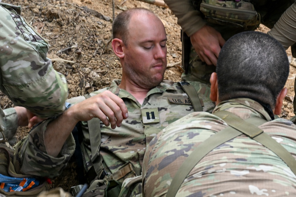 A U.S. Army Captain is being assisted by multiple soldiers in what appears to be a field medical training or casualty evacuation exercise in an outdoor, rocky terrain environment. The central figure wears U.S. Army OCP (Operational Camouflage Pattern) uniform with Captain rank insignia visible on his chest. Multiple personnel are attending to him, adjusting straps or harnesses, suggesting a simulated casualty scenario or extraction drill.
