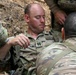 A U.S. Army Captain is being assisted by multiple soldiers in what appears to be a field medical training or casualty evacuation exercise in an outdoor, rocky terrain environment. The central figure wears U.S. Army OCP (Operational Camouflage Pattern) uniform with Captain rank insignia visible on his chest. Multiple personnel are attending to him, adjusting straps or harnesses, suggesting a simulated casualty scenario or extraction drill.