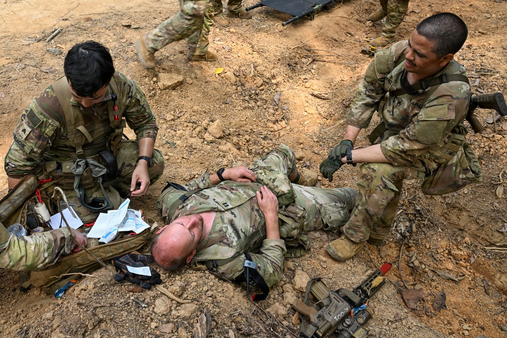 Two soldiers in MultiCam/OCP camouflage uniforms are performing tactical combat casualty care (TCCC) on a third soldier lying on the ground. The scene appears to be a field training exercise, with medical supplies scattered nearby and a rifle on the ground. A fourth soldier is visible in the background walking away.