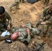 Two soldiers in MultiCam/OCP camouflage uniforms are performing tactical combat casualty care (TCCC) on a third soldier lying on the ground. The scene appears to be a field training exercise, with medical supplies scattered nearby and a rifle on the ground. A fourth soldier is visible in the background walking away.