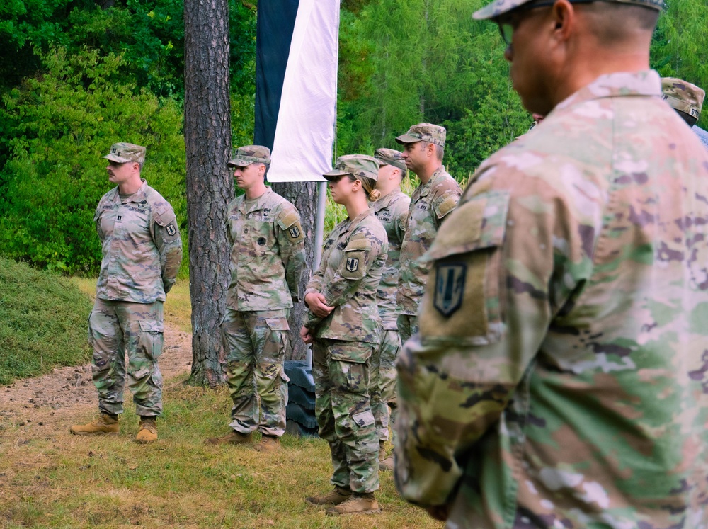 A group of U.S. Army soldiers in Operational Camouflage Pattern (OCP) uniforms are assembled outdoors in a wooded area, appearing to be at a formal military ceremony or formation. A white flag or banner is partially visible hanging from a tree in the background. The soldiers are standing at attention or in a relaxed parade rest posture.