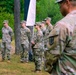 A group of U.S. Army soldiers in Operational Camouflage Pattern (OCP) uniforms are assembled outdoors in a wooded area, appearing to be at a formal military ceremony or formation. A white flag or banner is partially visible hanging from a tree in the background. The soldiers are standing at attention or in a relaxed parade rest posture.