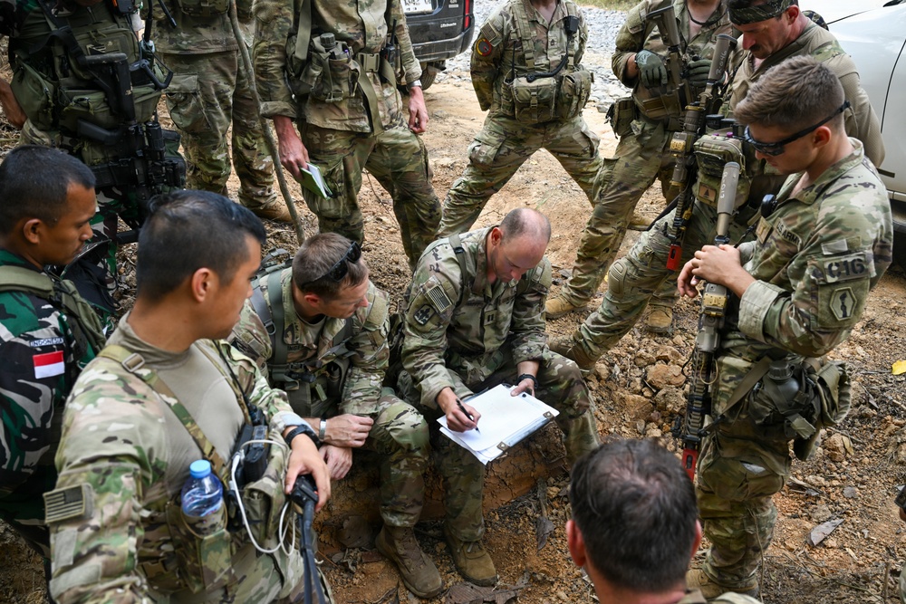A group of military personnel from at least two nations are gathered in an outdoor field setting, appearing to conduct a briefing or planning session. Two soldiers are crouched on the ground reviewing documents on a clipboard while others stand around them. The presence of both US Army patches and an Indonesian flag patch suggests this is a joint multinational military exercise or operation.