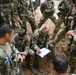 A group of military personnel from at least two nations are gathered in an outdoor field setting, appearing to conduct a briefing or planning session. Two soldiers are crouched on the ground reviewing documents on a clipboard while others stand around them. The presence of both US Army patches and an Indonesian flag patch suggests this is a joint multinational military exercise or operation.