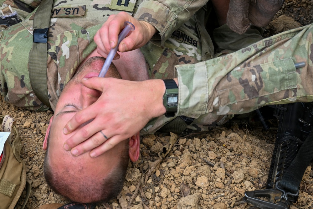 A U.S. Army soldier is lying on the ground receiving medical treatment from another soldier, likely during a field training exercise or combat scenario. A medic or fellow soldier is administering what appears to be eye drops or a nasal airway adjunct near the casualty's face. Both individuals are wearing OCP (Operational Camouflage Pattern) uniforms.