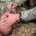 A U.S. Army soldier is lying on the ground receiving medical treatment from another soldier, likely during a field training exercise or combat scenario. A medic or fellow soldier is administering what appears to be eye drops or a nasal airway adjunct near the casualty's face. Both individuals are wearing OCP (Operational Camouflage Pattern) uniforms.