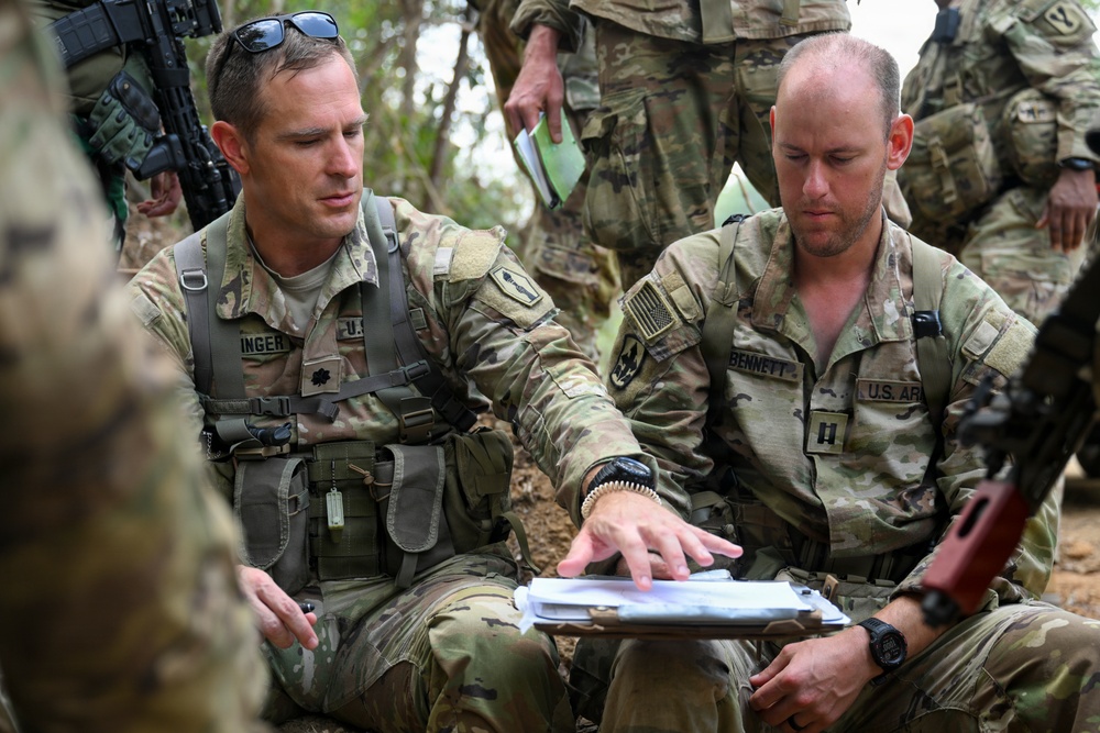 Two U.S. Army soldiers in MultiCam/OCP camouflage uniforms are reviewing documents on a clipboard or tablet in an outdoor field environment. The soldier on the left, with a partially visible name tag reading 'INGER' and a Lieutenant Colonel oak leaf rank insignia, is pointing at the documents while briefing the soldier on the right, identified as 'BENNETT' with a Captain rank insignia. Additional soldiers are visible in the background, suggesting a tactical planning or after-action review session.