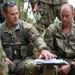 Two U.S. Army soldiers in MultiCam/OCP camouflage uniforms are reviewing documents on a clipboard or tablet in an outdoor field environment. The soldier on the left, with a partially visible name tag reading 'INGER' and a Lieutenant Colonel oak leaf rank insignia, is pointing at the documents while briefing the soldier on the right, identified as 'BENNETT' with a Captain rank insignia. Additional soldiers are visible in the background, suggesting a tactical planning or after-action review session.