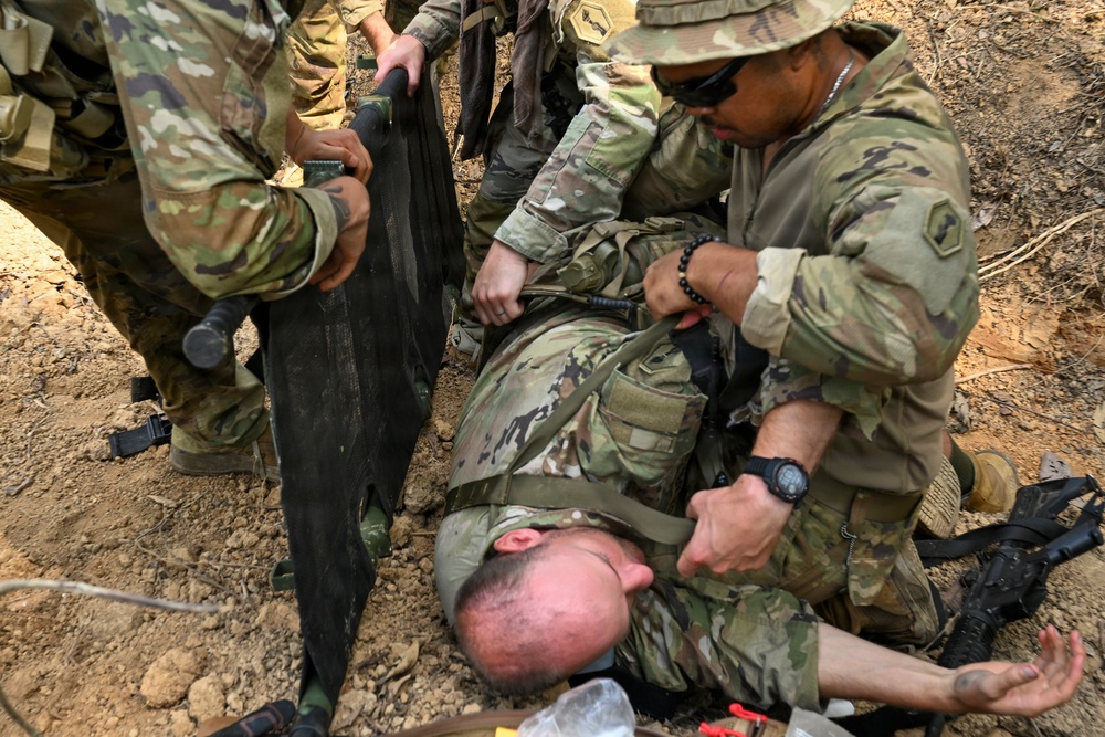 Multiple soldiers in OCP (Operational Camouflage Pattern) uniforms are performing a casualty evacuation or medical treatment drill on a downed soldier lying on the ground. Two standing/kneeling soldiers are positioning what appears to be a litter or stretcher beneath the casualty. The setting is an outdoor dirt terrain consistent with a field training exercise.