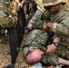 Multiple soldiers in OCP (Operational Camouflage Pattern) uniforms are performing a casualty evacuation or medical treatment drill on a downed soldier lying on the ground. Two standing/kneeling soldiers are positioning what appears to be a litter or stretcher beneath the casualty. The setting is an outdoor dirt terrain consistent with a field training exercise.