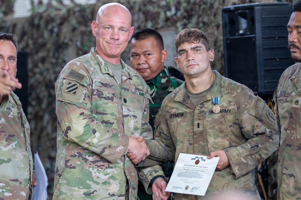 A U.S. Army award ceremony is taking place, with a senior officer presenting the Army Achievement Medal to a soldier named Efymow. The recipient is holding a Department of the Army certificate for the Army Achievement Medal and has the medal pinned to his uniform. The ceremony appears to be held in a military facility decorated with camouflage netting, with personnel from at least two different military forces present.