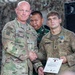 A U.S. Army award ceremony is taking place, with a senior officer presenting the Army Achievement Medal to a soldier named Efymow. The recipient is holding a Department of the Army certificate for the Army Achievement Medal and has the medal pinned to his uniform. The ceremony appears to be held in a military facility decorated with camouflage netting, with personnel from at least two different military forces present.