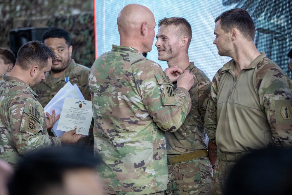 A military awards ceremony is taking place, with a senior soldier pinning a medal or award onto a junior soldier's uniform. A soldier to the left holds official Department of the Army paperwork, likely the award citation. Several other soldiers in U.S. Army OCP (Operational Camouflage Pattern) uniforms are present as witnesses.