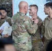 A military awards ceremony is taking place, with a senior soldier pinning a medal or award onto a junior soldier's uniform. A soldier to the left holds official Department of the Army paperwork, likely the award citation. Several other soldiers in U.S. Army OCP (Operational Camouflage Pattern) uniforms are present as witnesses.