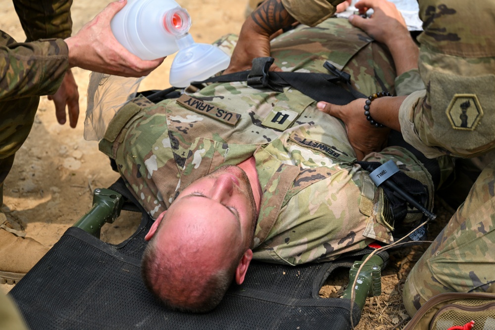 A U.S. Army soldier identified by the name tape 'BENNETT' is lying on a litter/stretcher and receiving emergency medical treatment. Multiple personnel are administering care, including bag-valve mask (BVM) ventilation and what appears to be tourniquet application. This appears to be either a combat casualty care scenario or a field medical training exercise.