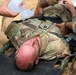 A U.S. Army soldier identified by the name tape 'BENNETT' is lying on a litter/stretcher and receiving emergency medical treatment. Multiple personnel are administering care, including bag-valve mask (BVM) ventilation and what appears to be tourniquet application. This appears to be either a combat casualty care scenario or a field medical training exercise.