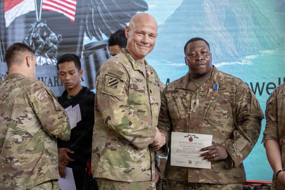 A U.S. Army awards ceremony is taking place, with a senior officer presenting a certificate and medal to a soldier identified by nametape as 'CADE'. The recipient is wearing an Army Achievement Medal on his uniform and holding a Department of the Army certificate. The event appears to be occurring at a joint or multinational military venue, evidenced by the presence of a civilian in the background wearing what appears to be Indonesian military attire.