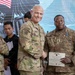 A U.S. Army awards ceremony is taking place, with a senior officer presenting a certificate and medal to a soldier identified by nametape as 'CADE'. The recipient is wearing an Army Achievement Medal on his uniform and holding a Department of the Army certificate. The event appears to be occurring at a joint or multinational military venue, evidenced by the presence of a civilian in the background wearing what appears to be Indonesian military attire.