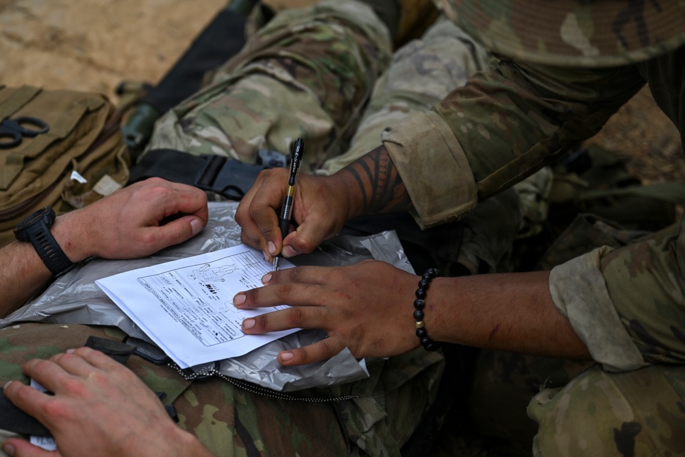 Two military personnel in MultiCam/OCP camouflage uniforms are filling out what appears to be a military medical or casualty documentation form. One individual is writing with a pen while the other holds the document steady on what appears to be a clear plastic sleeve or clipboard. The scene suggests a field training exercise or operational environment based on the sandy terrain visible in the background.
