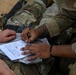 Two military personnel in MultiCam/OCP camouflage uniforms are filling out what appears to be a military medical or casualty documentation form. One individual is writing with a pen while the other holds the document steady on what appears to be a clear plastic sleeve or clipboard. The scene suggests a field training exercise or operational environment based on the sandy terrain visible in the background.