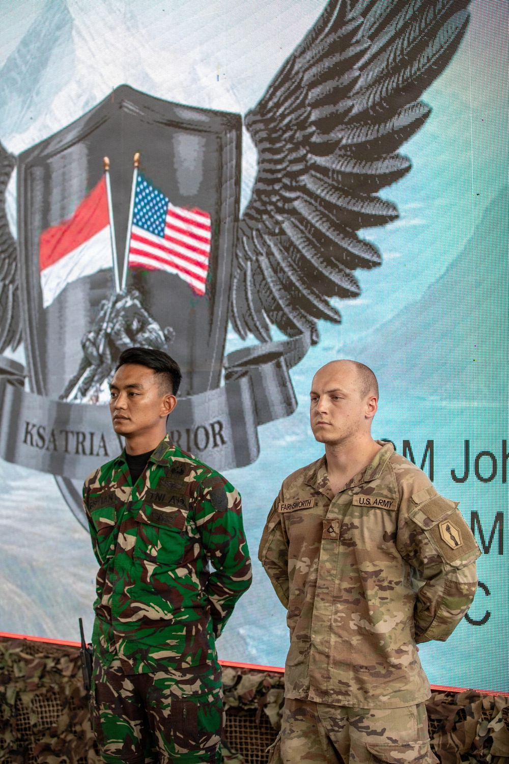 Two military personnel stand at attention in front of a large display banner showing crossed Indonesian and American flags with an eagle emblem and the text 'KSATRIA WARRIOR'. The soldier on the left wears Indonesian Army (TNI AD) camouflage uniform, while the soldier on the right wears U.S. Army OCP (MultiCam) uniform with the name tape 'FARNSWORTH' and a 1st Cavalry Division patch. This appears to be a joint military exercise or bilateral training event between Indonesia and the United States.