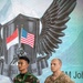 Two military personnel stand at attention in front of a large display banner showing crossed Indonesian and American flags with an eagle emblem and the text 'KSATRIA WARRIOR'. The soldier on the left wears Indonesian Army (TNI AD) camouflage uniform, while the soldier on the right wears U.S. Army OCP (MultiCam) uniform with the name tape 'FARNSWORTH' and a 1st Cavalry Division patch. This appears to be a joint military exercise or bilateral training event between Indonesia and the United States.