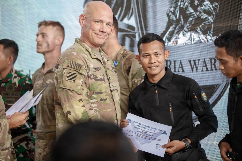 A U.S. Army soldier in MultiCam/OCP uniform is presenting a certificate from the Joint Pacific Multinational Readiness Center (JPMRC) to a recipient wearing a black tactical uniform. The ceremony appears to be a multinational military training exercise or event, with personnel from multiple nations visible in the background, including what appears to be Indonesian military (TNI AD) soldiers. Both the presenter and recipient are smiling for the photo.
