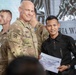 A U.S. Army soldier in MultiCam/OCP uniform is presenting a certificate from the Joint Pacific Multinational Readiness Center (JPMRC) to a recipient wearing a black tactical uniform. The ceremony appears to be a multinational military training exercise or event, with personnel from multiple nations visible in the background, including what appears to be Indonesian military (TNI AD) soldiers. Both the presenter and recipient are smiling for the photo.