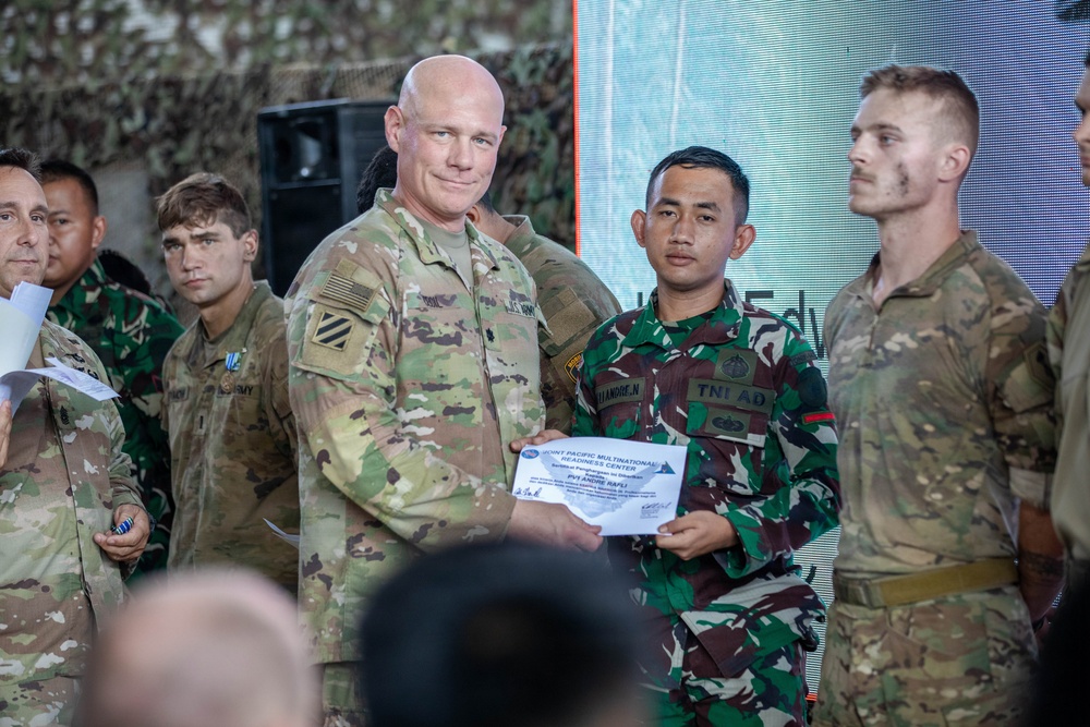 A U.S. Army officer is presenting a certificate from the Joint Pacific Multinational Readiness Center to an Indonesian Army (TNI AD) soldier during what appears to be a multinational military exercise or training event. Multiple soldiers from different nations are visible in the background, suggesting a joint military cooperation ceremony. The setting appears to be an outdoor or semi-covered venue decorated with camouflage netting.