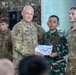 A U.S. Army officer is presenting a certificate from the Joint Pacific Multinational Readiness Center to an Indonesian Army (TNI AD) soldier during what appears to be a multinational military exercise or training event. Multiple soldiers from different nations are visible in the background, suggesting a joint military cooperation ceremony. The setting appears to be an outdoor or semi-covered venue decorated with camouflage netting.