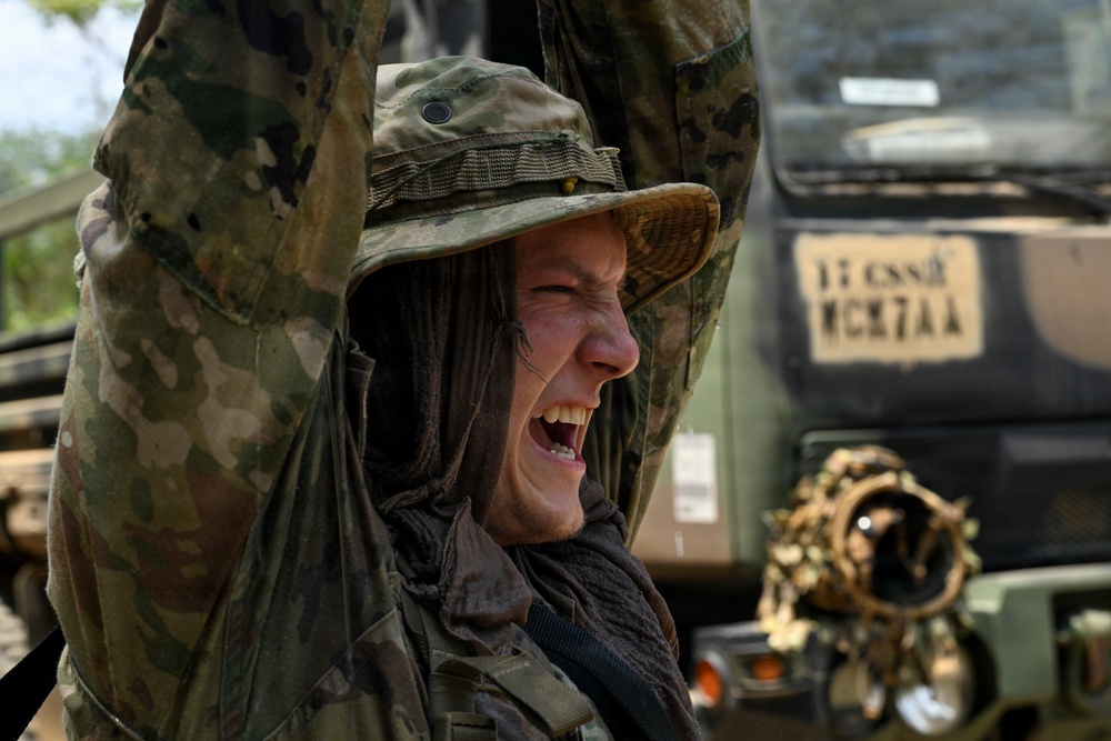 A soldier in full camouflage uniform and boonie hat is laughing or reacting expressively with arms raised, appearing to be in a field environment. Military vehicles are visible in the background, suggesting an active training exercise or operational deployment. The soldier is wearing multicam pattern gear including a shemagh neck covering and tactical equipment.