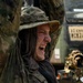 A soldier in full camouflage uniform and boonie hat is laughing or reacting expressively with arms raised, appearing to be in a field environment. Military vehicles are visible in the background, suggesting an active training exercise or operational deployment. The soldier is wearing multicam pattern gear including a shemagh neck covering and tactical equipment.