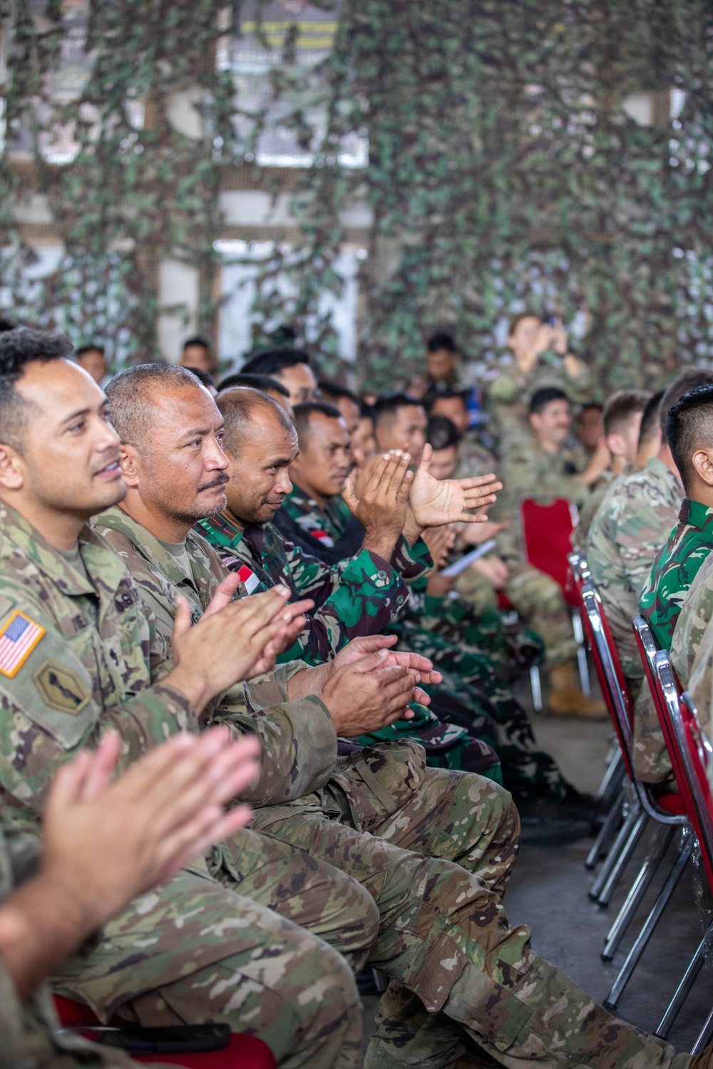 A group of military personnel from at least two nations are seated in rows and applauding during what appears to be a joint military ceremony or briefing. Visible uniforms include US Army OCP (Operational Camouflage Pattern) with American flag patches and Indonesian Army (TNI-AD) woodland camouflage uniforms with red-and-white Indonesian flag patches. The venue is decorated with camouflage netting overhead.