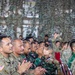 A group of military personnel from at least two nations are seated in rows and applauding during what appears to be a joint military ceremony or briefing. Visible uniforms include US Army OCP (Operational Camouflage Pattern) with American flag patches and Indonesian Army (TNI-AD) woodland camouflage uniforms with red-and-white Indonesian flag patches. The venue is decorated with camouflage netting overhead.