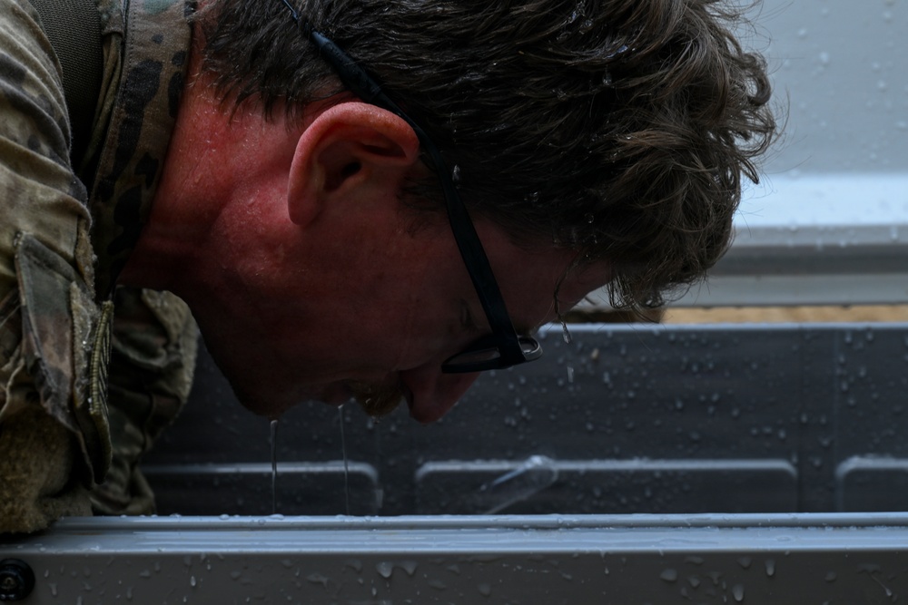 A person wearing military camouflage uniform and black-framed glasses is leaning forward with their face close to a wet metal surface. Water droplets are visibly dripping from their face and glasses, suggesting recent exposure to rain or water immersion. The close-up shot emphasizes physical exertion or exhaustion in wet conditions.