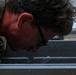 A person wearing military camouflage uniform and black-framed glasses is leaning forward with their face close to a wet metal surface. Water droplets are visibly dripping from their face and glasses, suggesting recent exposure to rain or water immersion. The close-up shot emphasizes physical exertion or exhaustion in wet conditions.