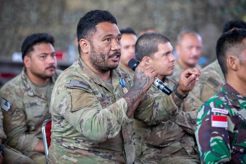 A group of military personnel from multiple nations are seated together, likely during a briefing or discussion session. The central figure, wearing US Army multicam uniform with a medal and visible arm tattoos, is speaking into a microphone. An Indonesian military soldier is visible in the foreground right, identifiable by the Indonesia patch and distinctive camouflage pattern.