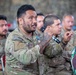 A group of military personnel from multiple nations are seated together, likely during a briefing or discussion session. The central figure, wearing US Army multicam uniform with a medal and visible arm tattoos, is speaking into a microphone. An Indonesian military soldier is visible in the foreground right, identifiable by the Indonesia patch and distinctive camouflage pattern.