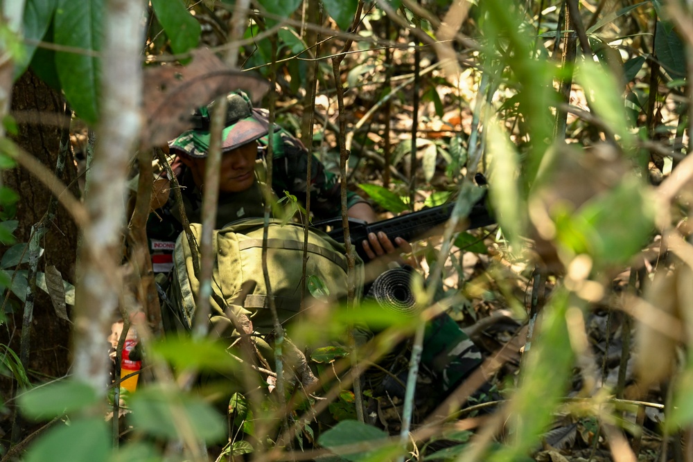 A soldier in Indonesian military camouflage uniform is concealed within dense jungle vegetation, holding a firearm. The individual is wearing a camouflage hat and has a patch visible on the sleeve that reads 'INDONESIA'. The scene appears to depict a jungle warfare training exercise or tactical concealment position.